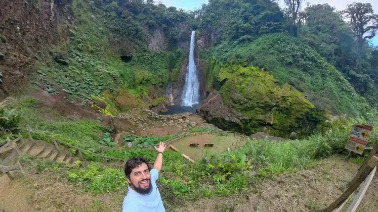 Un hombre sonriente con barba se toma una selfie mientras señala una cascada alta en un valle exuberante y verde.