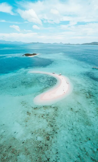 Vue aérienne d'un voyage en groupe WeRoad sur un banc de sable rose en forme de croissant dans des eaux turquoise, avec des bateaux et des îles lointaines.