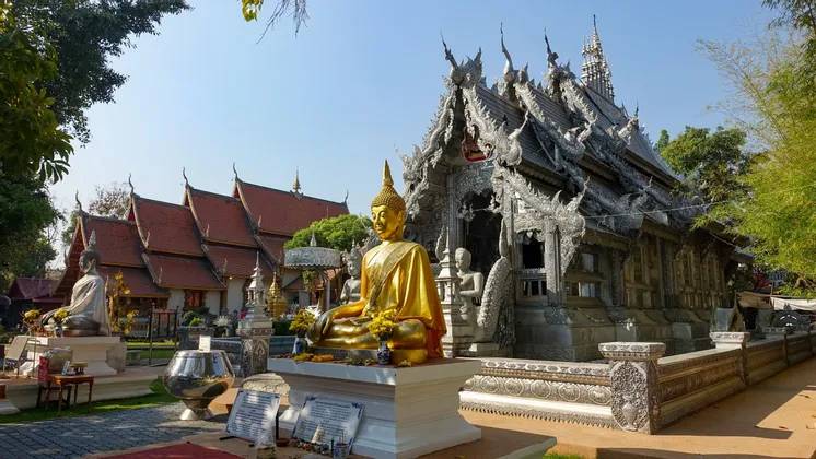 Eine goldene Buddha-Statue steht in einem Tempelhof vor einem reich verzierten Silbertempel und einem weiteren mit rotem Ziegeldach.