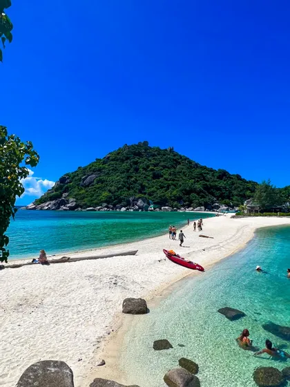 La gente camina por un banco de arena blanca que lleva a una exuberante isla verde, con un kayak rojo en la playa y nadadores en el agua cristalina color turquesa.