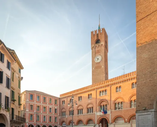 Una torre del reloj de ladrillo rojo iluminada por el sol y un edificio adyacente con arcos se alzan en una plaza histórica bajo un cielo azul pálido.