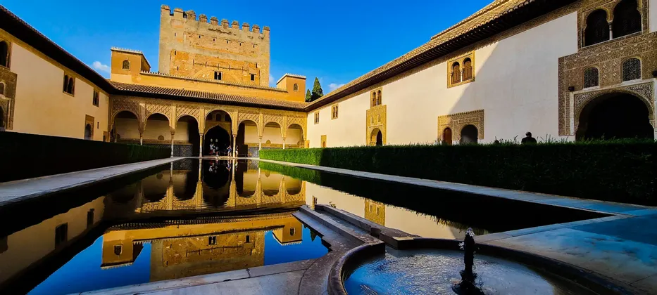 Un cortile in stile moresco con una lunga vasca riflettente che rispecchia gli edifici circostanti sotto un cielo azzurro limpido.