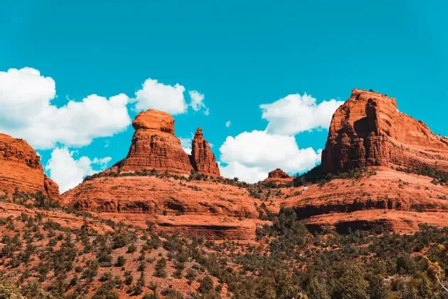 Grandi formazioni rocciose rosse si ergono da un paesaggio di macchia verde sotto un cielo azzurro brillante con nuvole bianche.