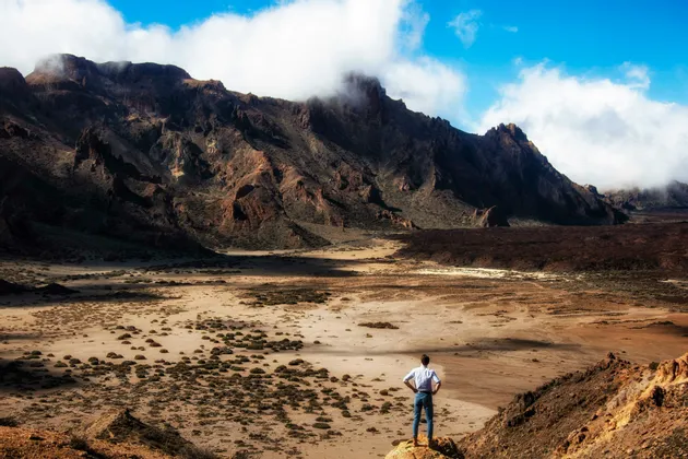 Un uomo in camicia bianca sta su uno sperone roccioso che domina una vasta valle arida con montagne aspre sullo sfondo.