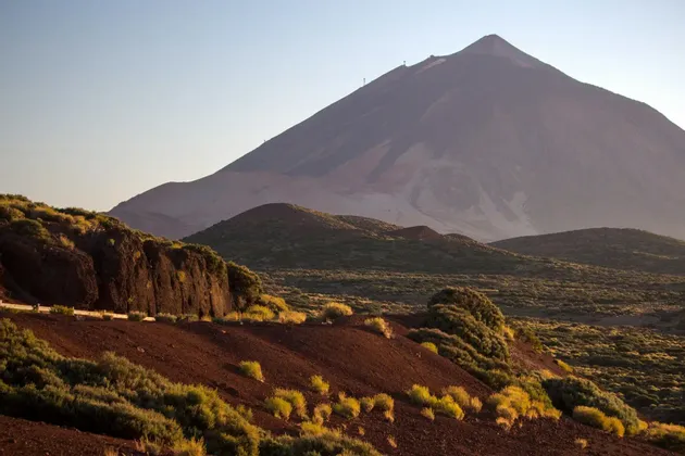 Un grande vulcano si erge su un paesaggio di terra vulcanica rossa e arbusti verdi sotto un cielo pallido e limpido.