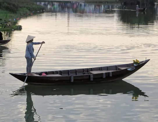 Una persona con sombrero cónico está de pie en un largo bote de madera, remando en aguas tranquilas y reflectantes con un solo remo.