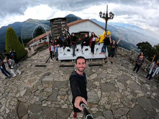 Un hombre se toma una selfie de gran angular con un viaje en grupo de WeRoad posando alrededor de un letrero de 'BOGOTÁ' en una plaza de piedra.