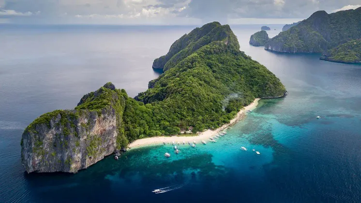Luftaufnahme einer üppig grünen Tropeninsel mit einem weißen Sandstrand und Booten, die im klaren türkisfarbenen Wasser vertäut sind.