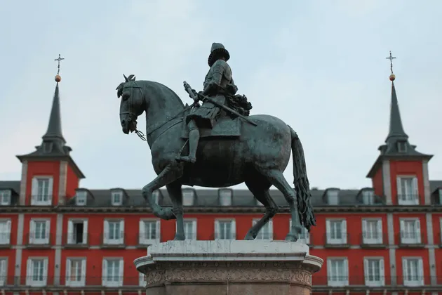 Une statue équestre en bronze sur un piédestal, avec un grand bâtiment rouge et ses tours jumelles en arrière-plan sous un ciel couvert.
