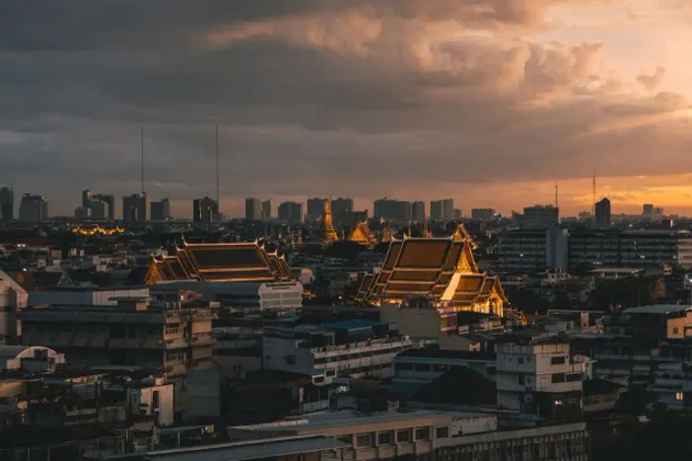Les toits de temples illuminés se détachent sur un paysage urbain dense avec une ligne d'horizon moderne au coucher du soleil sous un ciel nuageux.