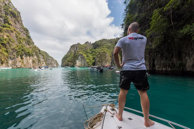Un hombre con una camiseta de WeRoad de pie en la proa de un barco, contemplando una laguna turquesa rodeada de acantilados rocosos.