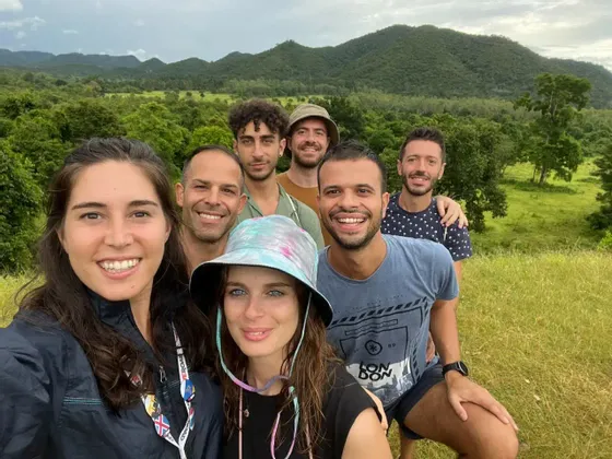 Un voyage en groupe WeRoad de sept personnes souriant pour un selfie dans un champ herbeux, avec en toile de fond des collines et des arbres verdoyants.