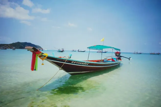 Ein traditionelles Longtail-Boot, mit Bändern geschmückt, gleitet in ruhigem, türkisfarbenem Wasser, mit einer grünen Insel am Horizont.