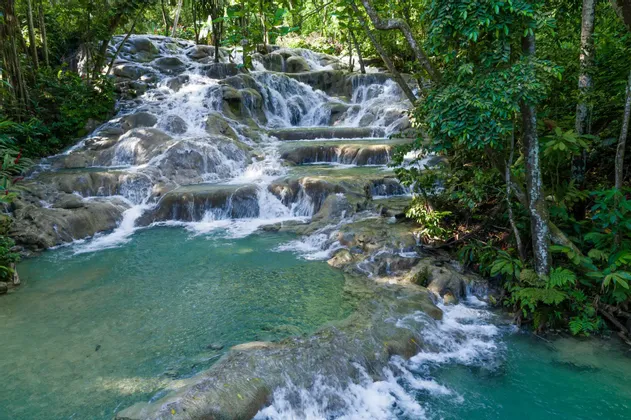 Ein terrassierter Wasserfall stürzt über Felsen in ein klares, türkisfarbenes Becken, umgeben von üppiger grüner Dschungelvegetation.