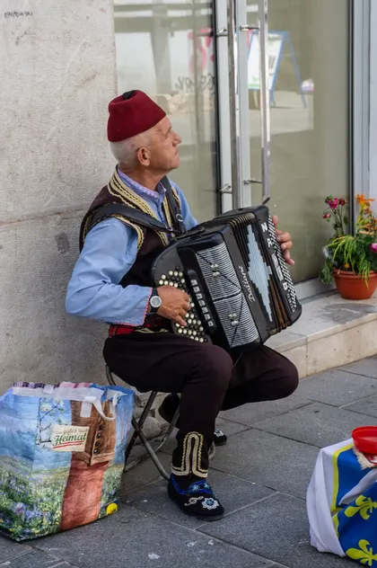 Un anciano con ropa tradicional, incluyendo un fez rojo y un chaleco bordado, está sentado en un taburete en una acera tocando el acordeón.