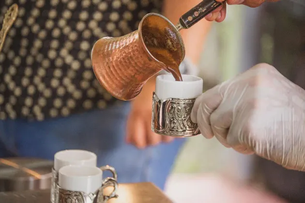 Una persona vierte café tradicional de una cafetera de cobre en una pequeña taza ornamentada sostenida por una mano enguantada.