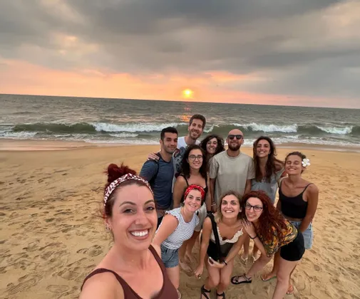 Un viaggio di gruppo WeRoad si fa un selfie su una spiaggia sabbiosa mentre il sole tramonta sull'oceano.
