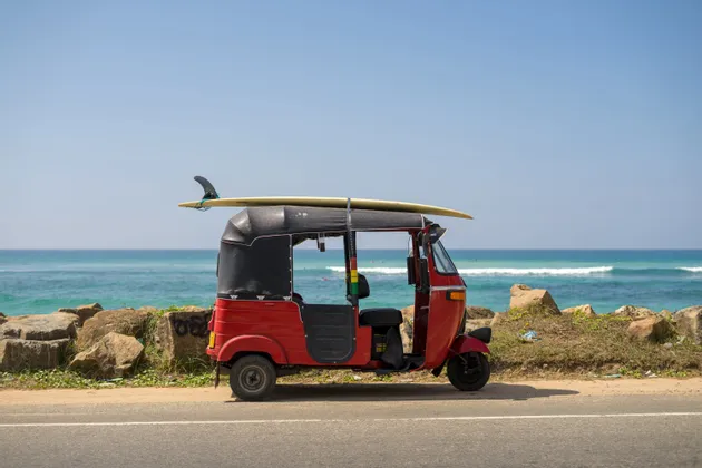 Un tuk-tuk rosso con una tavola da surf legata al tetto è parcheggiato su una strada costiera con l'oceano sullo sfondo.
