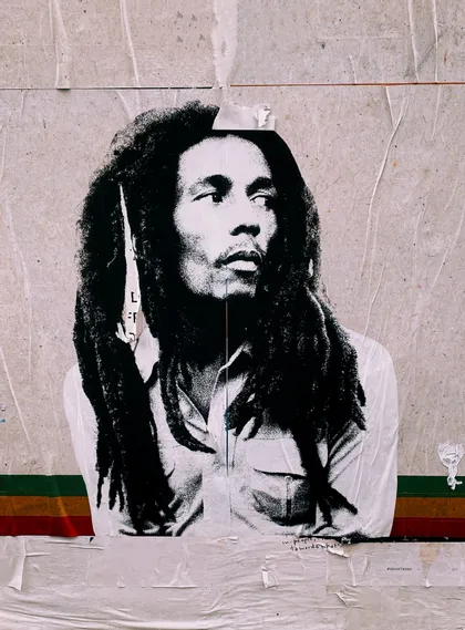 Ein Schwarz-Weiß-Paste-up-Plakat von Bob Marley mit Dreadlocks löst sich von einer strukturierten Wand über einem rot-gelb-grünen Streifen.