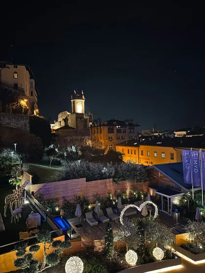 Una vista notturna di una terrazza d'albergo illuminata con statue di giraffa, che si affaccia su una città collinare con un antico castello.