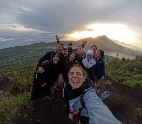 Un viaje en grupo de WeRoad se toma una selfie divertida con ojo de pez en la cima de una montaña al amanecer, con vistas a un volcán y un lago.