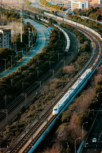 Veduta aerea di un treno proiettile bianco che viaggia su binari curvi, affiancato da un'autostrada tortuosa e altre linee ferroviarie.