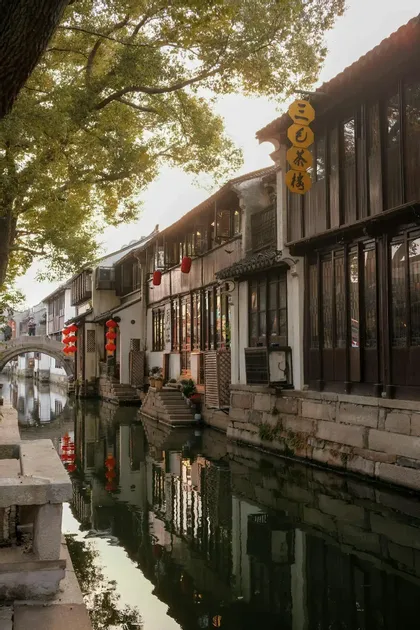 Edifici tradizionali con lanterne rosse costeggiano un canale calmo, con i loro riflessi che luccicano nell'acqua sotto un ponte in pietra ad arco.