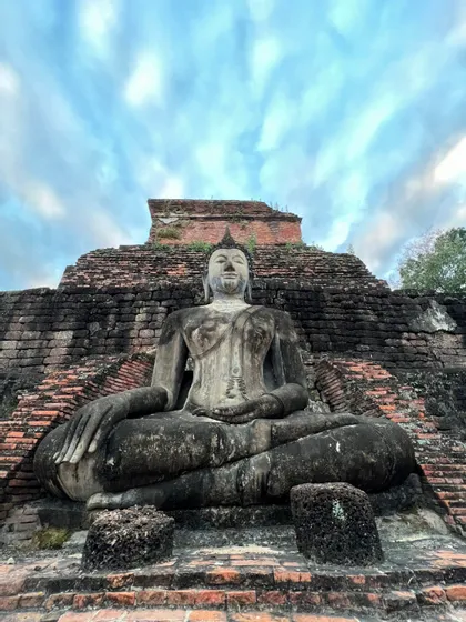 Una gran estatua de Buda de piedra erosionada se asienta en posición de meditación ante antiguas ruinas de ladrillo bajo un cielo nublado.
