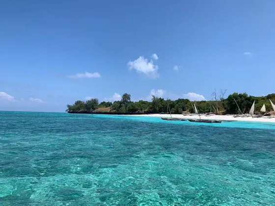 Acqua turchese cristallina in primo piano con barche a vela ormeggiate su una spiaggia di sabbia bianca, una costa alberata e un cielo blu.