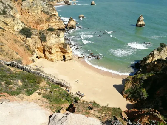 Una veduta dall'alto di una cala sabbiosa, circondata da scogliere gialle, con scale in legno che conducono all'oceano turchese e ai faraglioni.