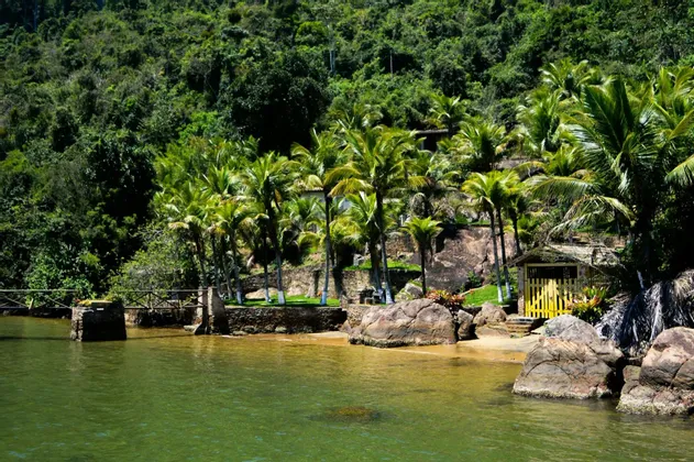Panorama di un'insenatura tropicale con piccola spiaggia sabbiosa, palme e una capanna, sullo sfondo una lussureggiante collina boscosa.