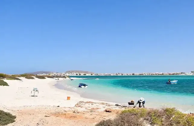 Una persona con un sombrero blanco haciendo una barbacoa en una playa de arena blanca junto a agua turquesa, con un pueblo visible al otro lado de la bahía.