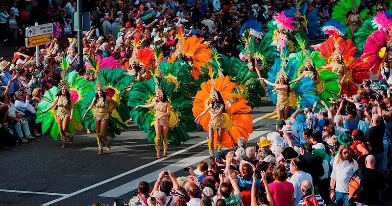 Danzatori in costumi colorati e piumati sfilano per una strada, osservati da una grande folla di spettatori.