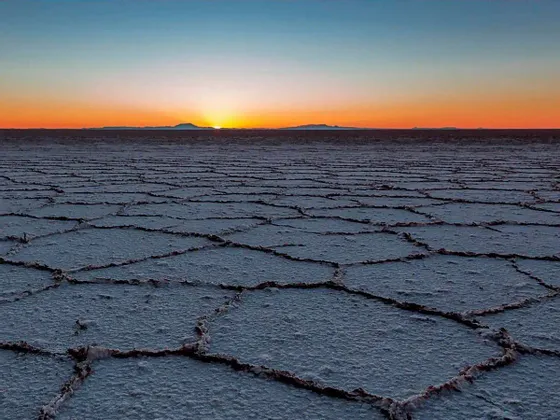 Il sole tramonta all'orizzonte su una vasta salina con un motivo geometrico e screpolato sul terreno.