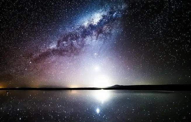 La Via Lattea e innumerevoli stelle nel cielo notturno si riflettono perfettamente su uno specchio d'acqua calmo all'orizzonte.
