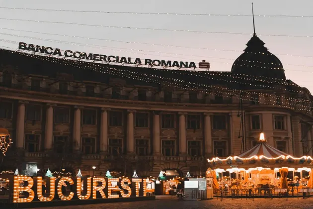 Una piazza cittadina al crepuscolo con un'insegna illuminata 'Bucuresti', una giostra e un grande edificio decorato con luci festive.