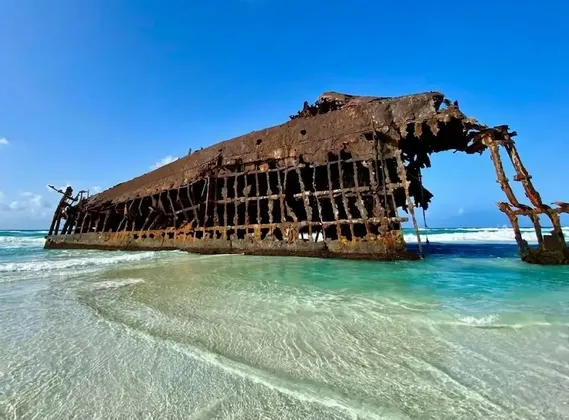 Un gran naufragio oxidado descansa en una playa de arena, parcialmente sumergido en aguas cristalinas de color turquesa bajo un cielo azul.