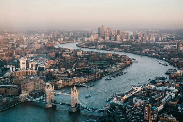 An aerial view of Tower Bridge and the River Thames winding through a dense cityscape towards a modern skyline under a hazy sky.