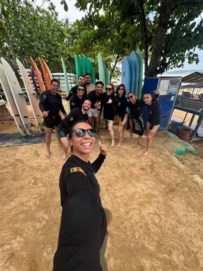 Un groupe WeRoad se prend en selfie sur une plage de sable devant des râteliers de planches de surf colorées.