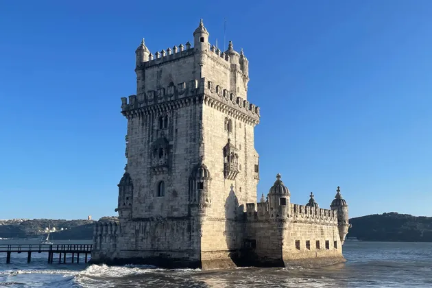 An ornate, fortified stone tower stands in the water under a clear blue sky, with gentle waves lapping at its base.
