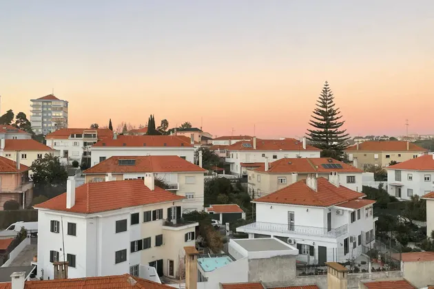 A cluster of white houses with red-tiled roofs on a hillside under a warm, pastel-colored sunset sky.