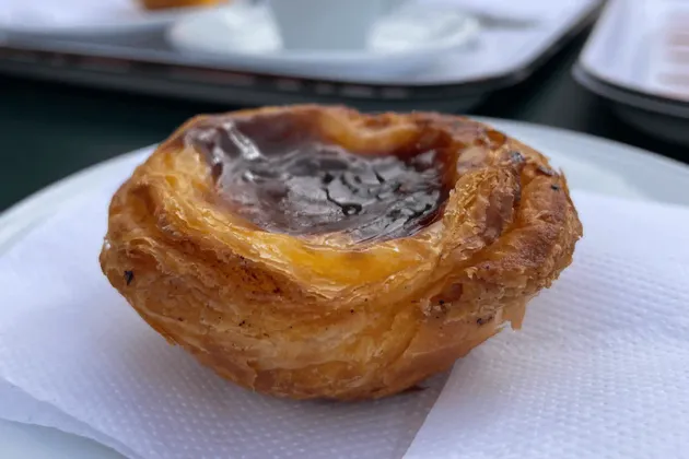 A close-up of a pastel de nata, a Portuguese egg tart, with a flaky crust and caramelized top, sitting on a white napkin.