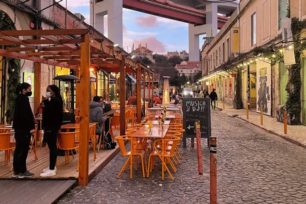 People dining at an outdoor restaurant with orange chairs on a cobblestone street under a large bridge at dusk.
