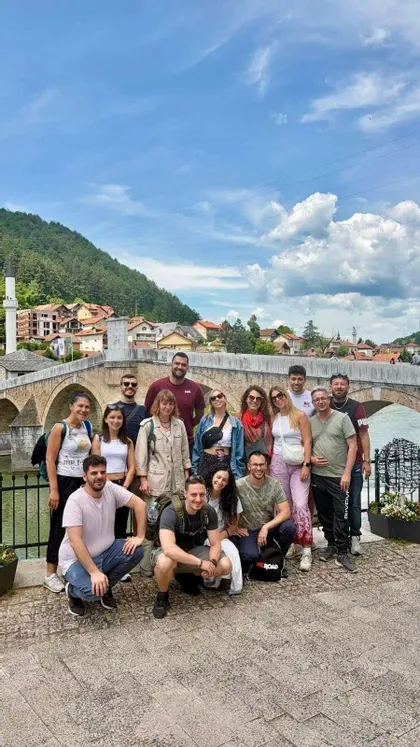 Un viaggio di gruppo WeRoad posa per una foto di fronte a un grande ponte di pietra, con una città e una collina verde sullo sfondo.