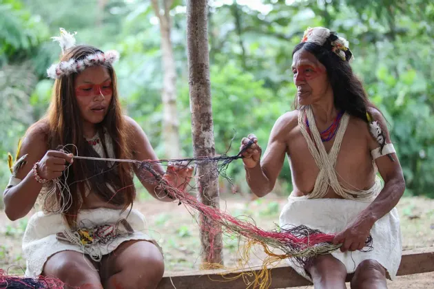 Dos mujeres indígenas con ropa tradicional y tocados de plumas trabajan juntas con hilos coloridos al aire libre