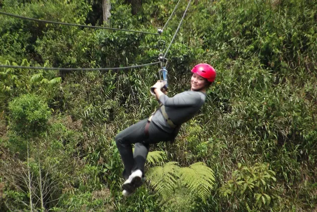 Una persona con casco rosso sorride mentre fa zipline in una fitta foresta verde.