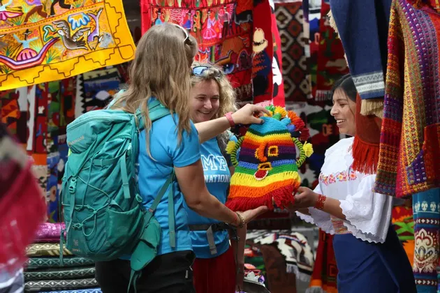Dos mujeres en un viaje grupal de WeRoad miran un artículo de punto colorido sostenido por un vendedor local en un mercado al aire libre.