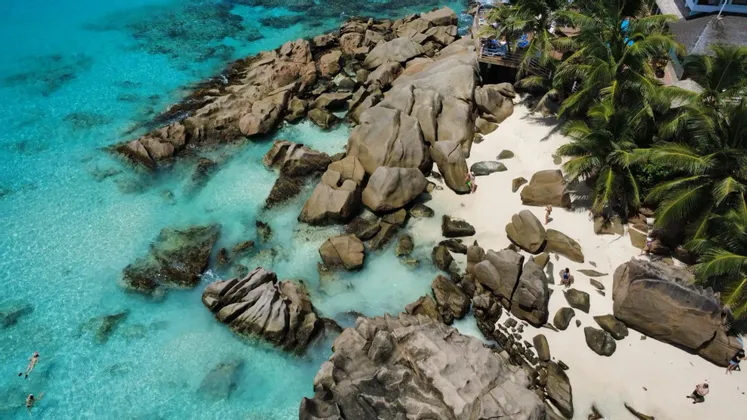 Veduta aerea di una costa rocciosa con una spiaggia di sabbia bianca, palme e persone che nuotano in acque cristalline turchesi.