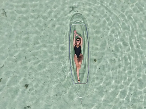 Vista dall'alto di una donna in costume nero sdraiata in un kayak trasparente che galleggia su acqua limpida e poco profonda.