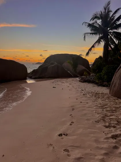Impronte nella sabbia su una spiaggia tropicale con grandi massi e una palma durante un tramonto colorato.